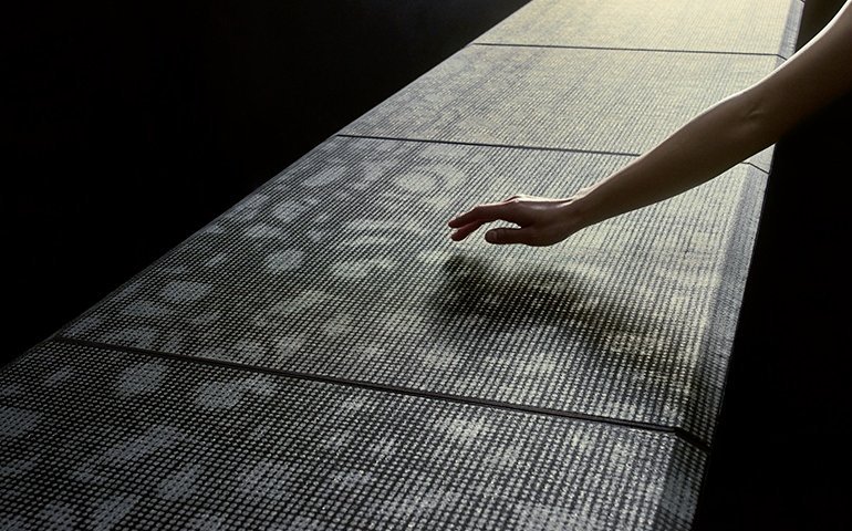 金属糸が織り込まれた天板の上に伸びる腕と手、薄明かりと影模様が映る 