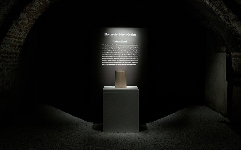 展示台に乗せられた円柱型の木製オブジェ、背景に説明文と見出し「Electronics Meets Crafts」  