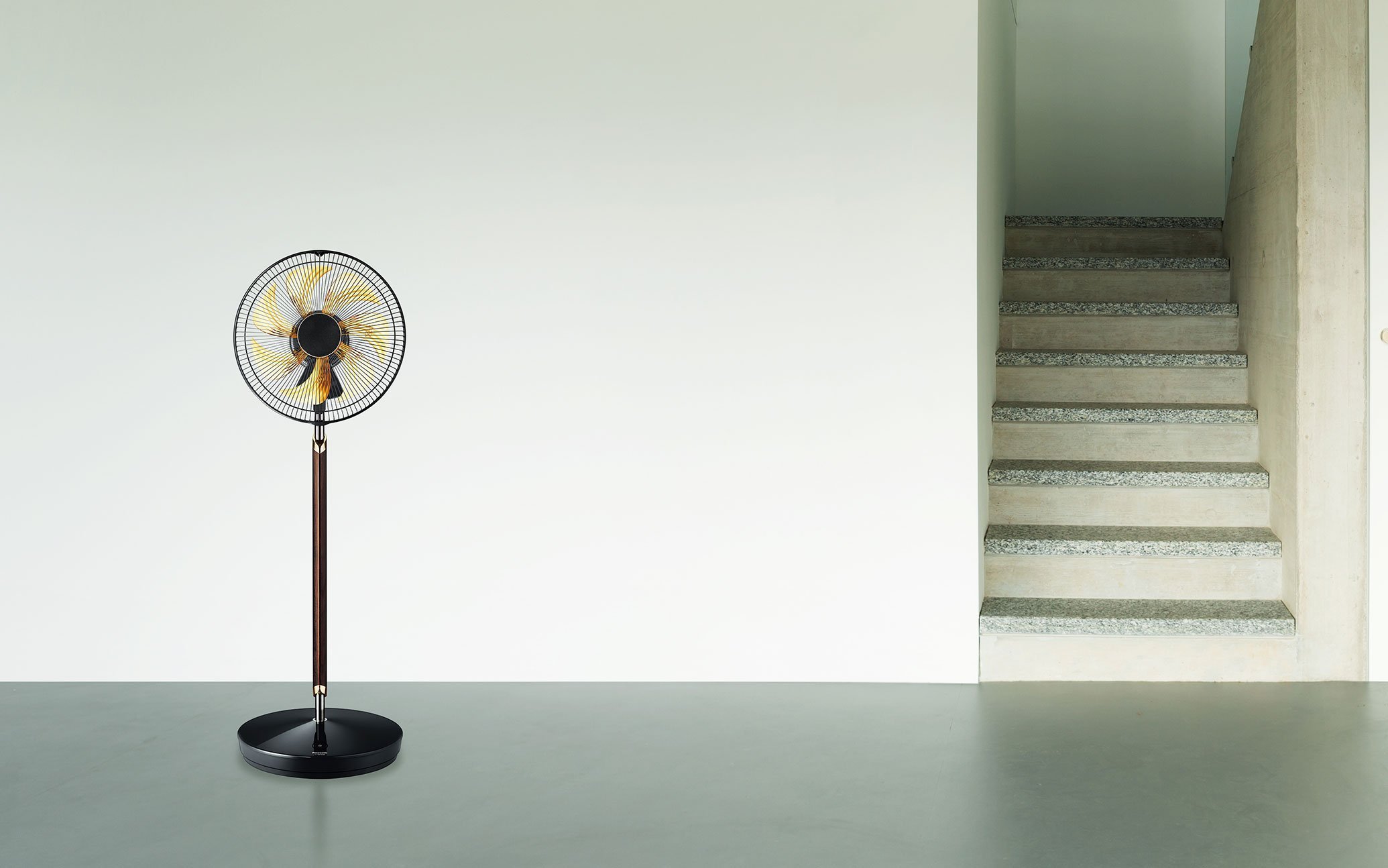 灰色の床に黄色い羽根を持つ黒色のスタンド扇風機が立っている。隣にはコンクリートの階段と白い壁が見える。