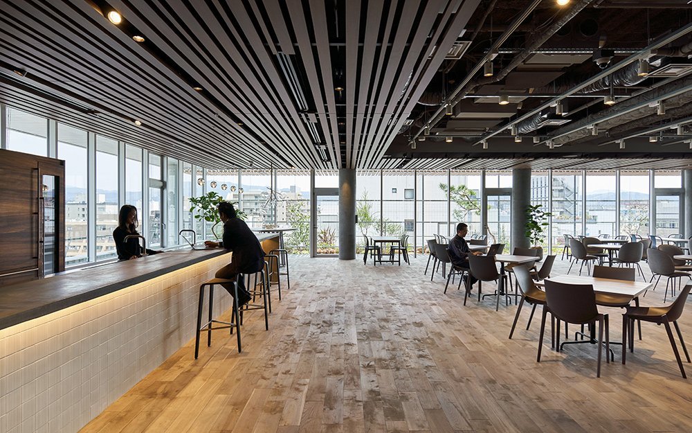 広い窓に囲まれた開放的な室内空間、木目調の床とカウンター、高い天井と複数のテーブルと椅子の配置