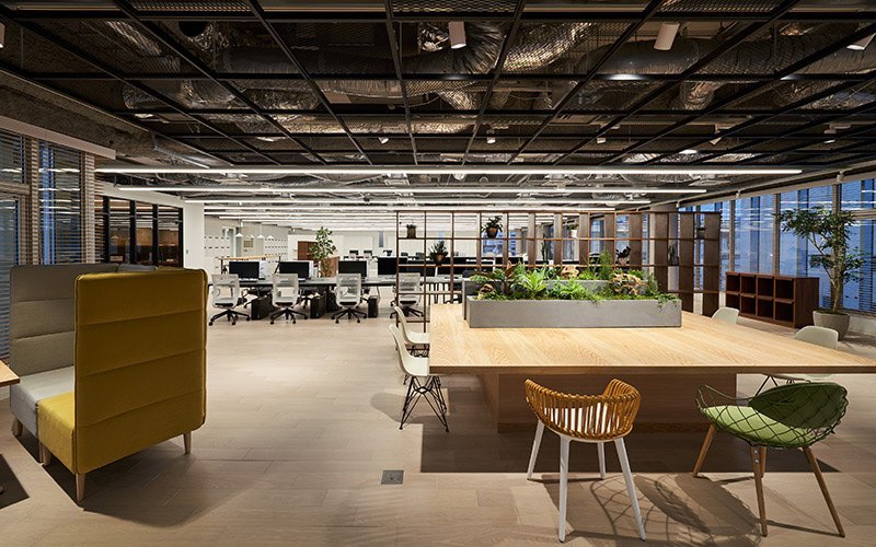 オープンな空間のオフィス、複数の作業デスクと会議用テーブルに植物と異なる椅子、照明と窓からの光