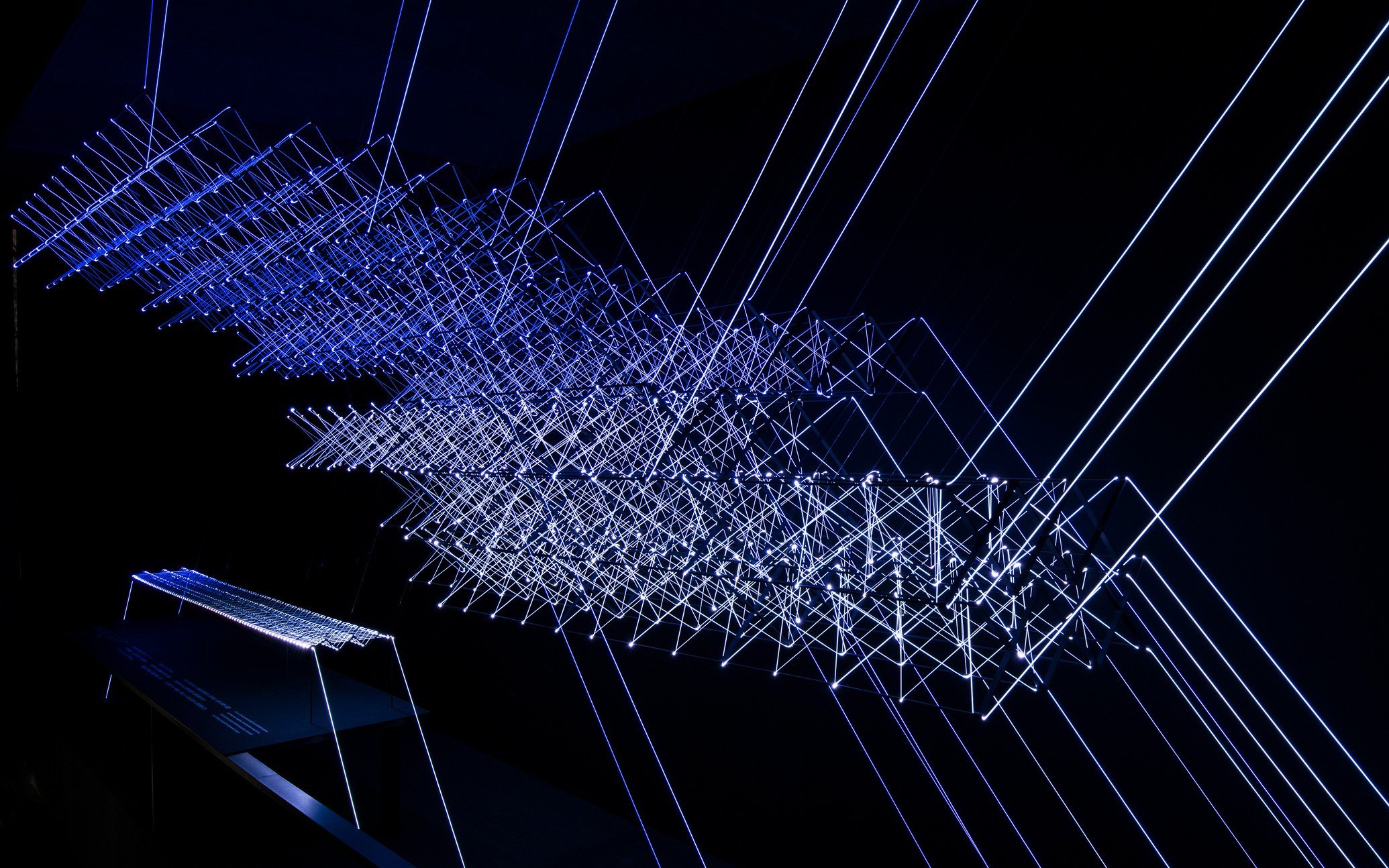 黒背景に青色の光線と格子状の構造を持つ立体的な光のインスタレーション