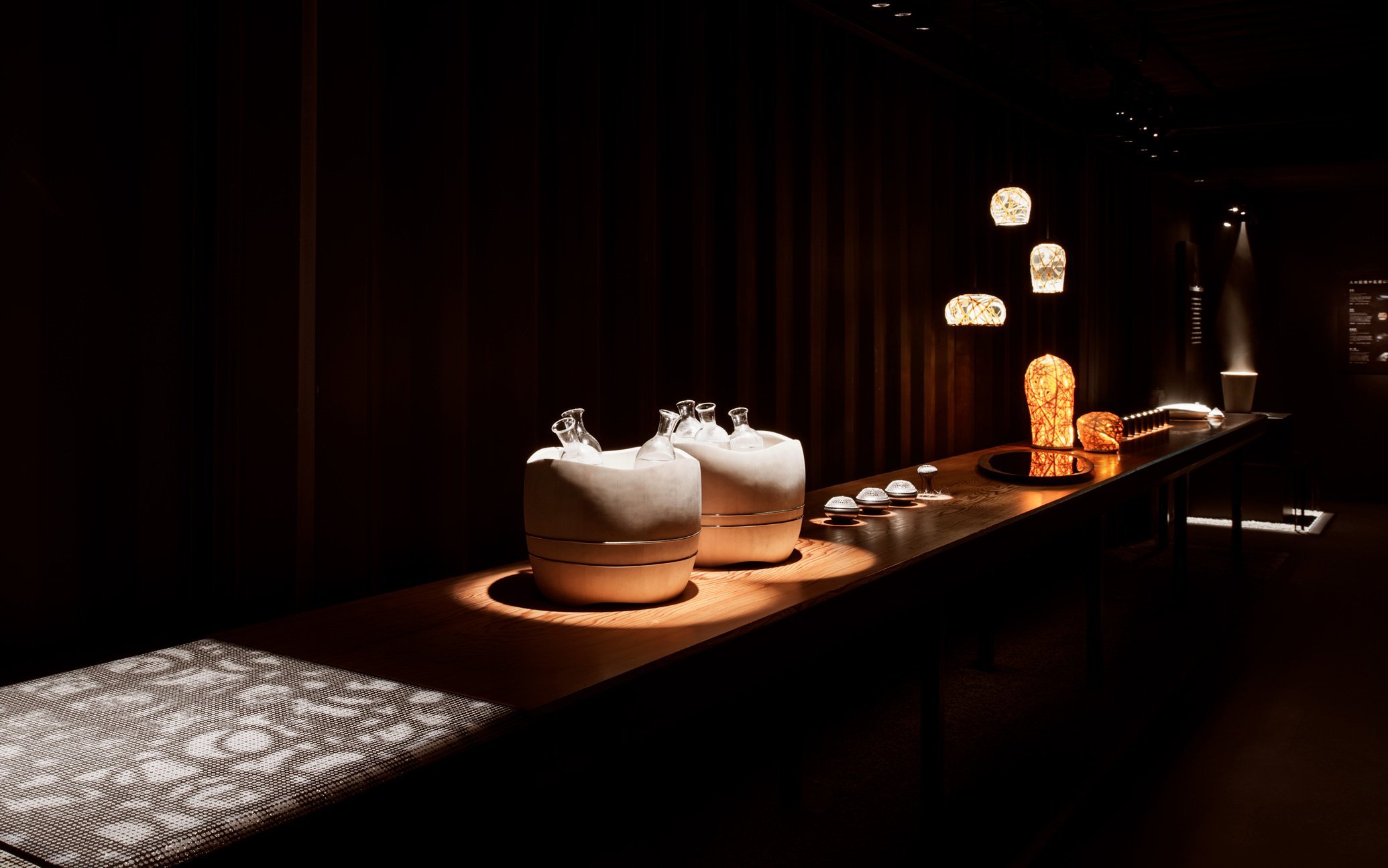 暗い展示室の長い木製テーブルにさまざまなデザインの照明器具とガラス容器のディスプレイ