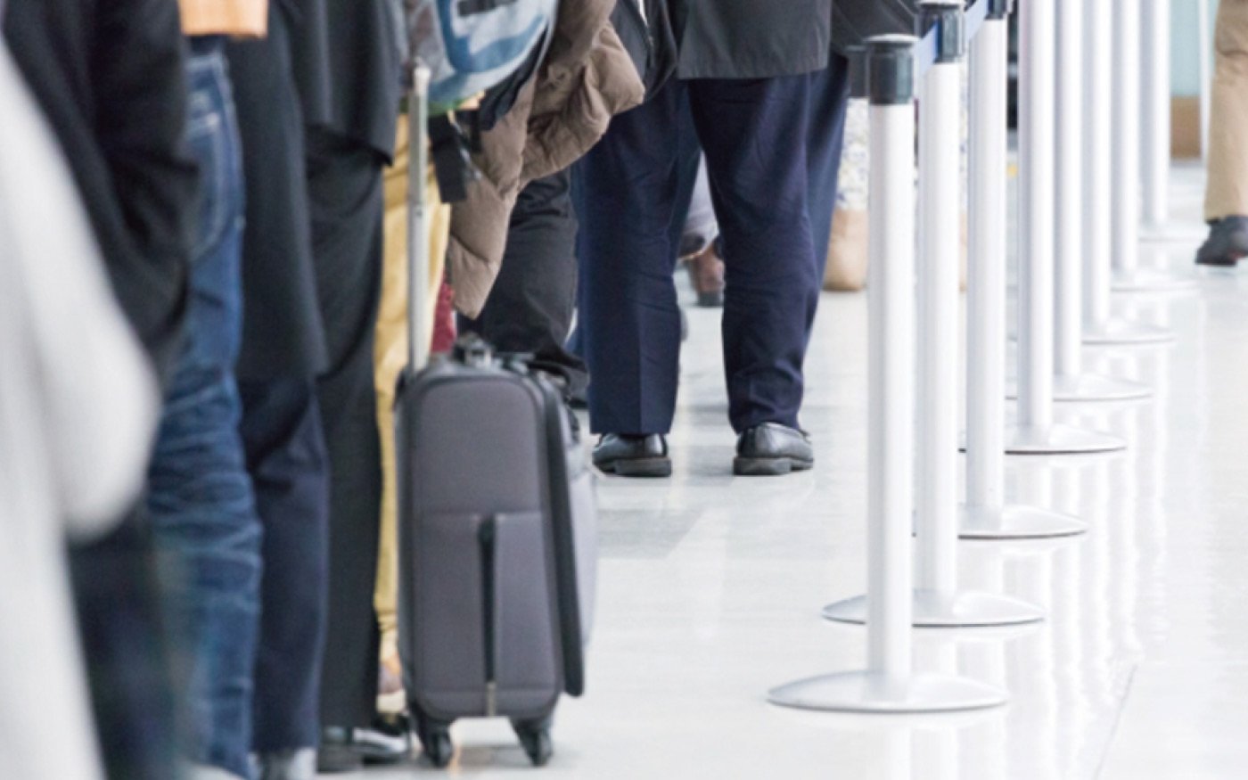 空港の列に並ぶ人々の脚元。スーツケースを引く人もいる。白い柱で区切られた通路と光沢のある床。
