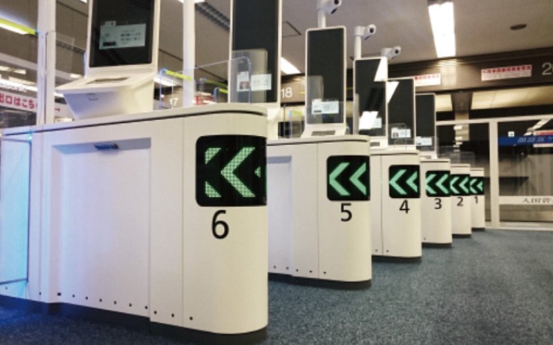 空港の入出国ゲートの白い端末が番号順に並ぶ。各端末に緑色の矢印表示と番号6から1まで。カーペット敷きの床と天井照明。