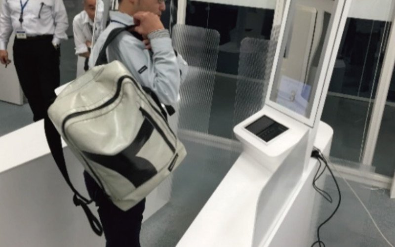  白い大きなリュックを背負った人物が透明なセルフチェックゲートの前に立つ。ゲートにはタッチパネルが設置され、周囲に複数の人がいる室内。