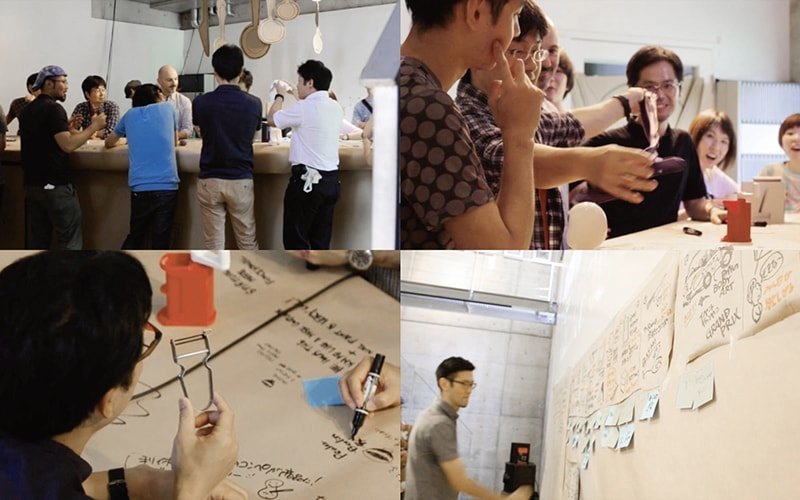 バーカウンターに並ぶ数名の男性と女性。打ち合わせ中のグループ、紙にメモや図を描く人、壁に貼られた付箋とメモでの作業空間。