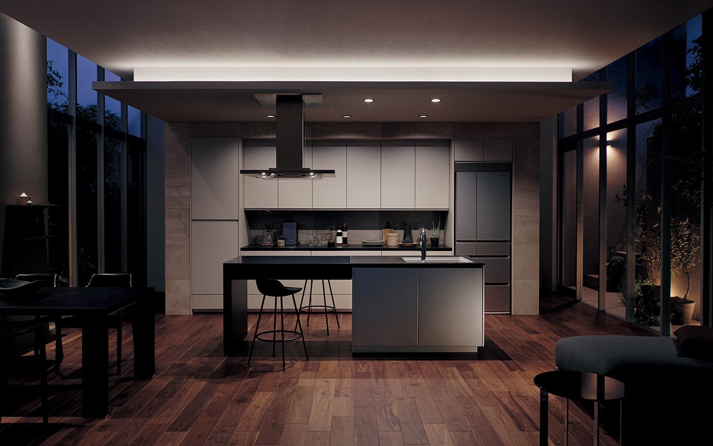 木目のフローリングに囲まれたモダンなグレーのキッチン。中央にアイランドカウンターとハイチェア、背面にキャビネットと冷蔵庫。ガラスの大きな窓と暖色の照明。