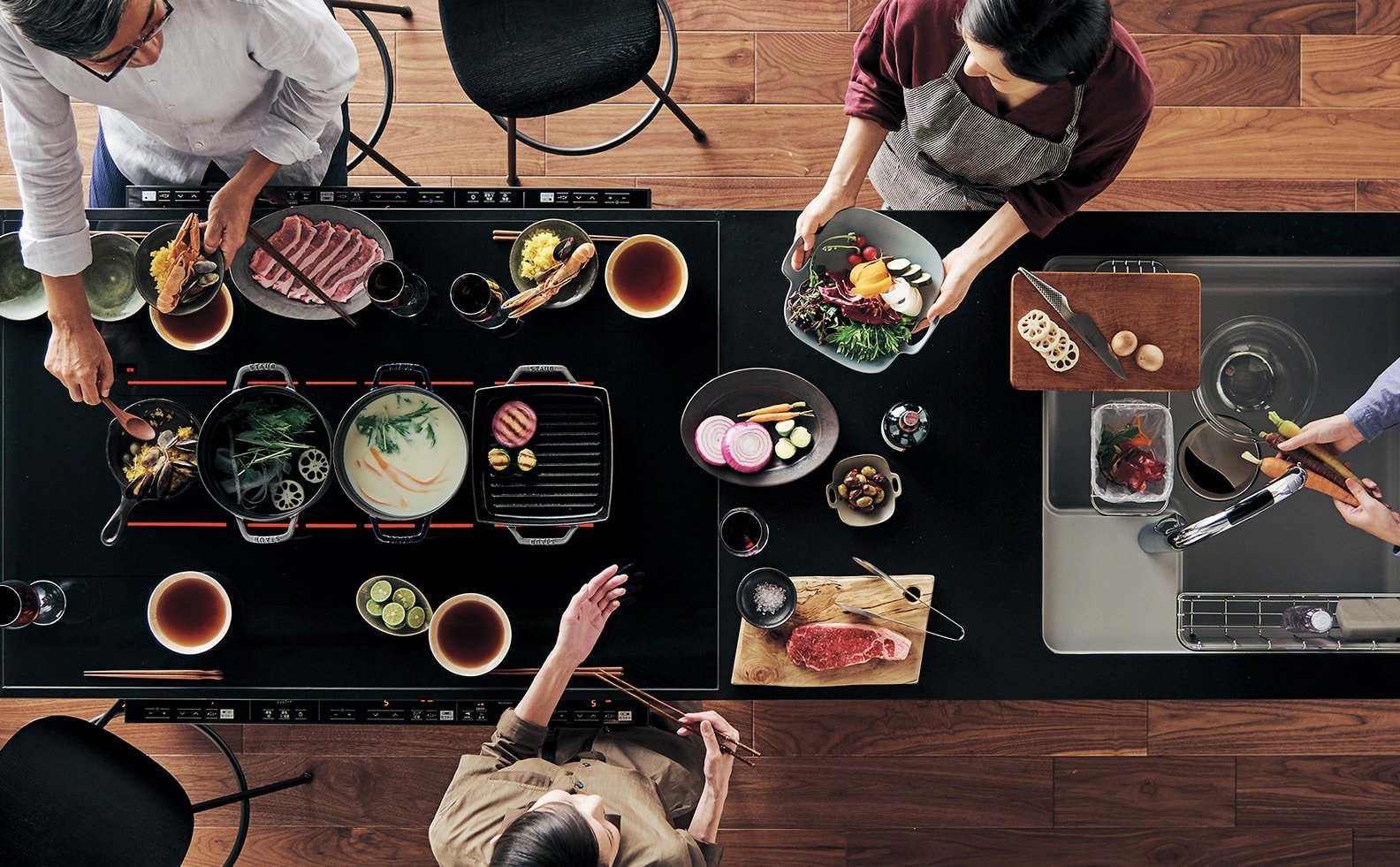 黒いキッチンカウンターの上に様々な料理が並んでいて、それを複数の人が取り囲んでいる