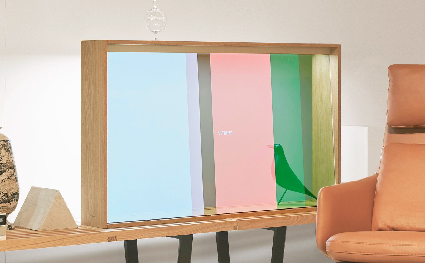 木製フレームの透明ディスプレイの前に緑色の鳥の置物と「VITRINE」の文字、側にオレンジ色の椅子