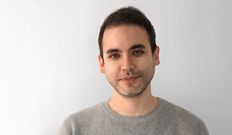 グレーのニットセーターを着た上半身。短い黒髪とひげ。白い背景の前でのポートレート撮影