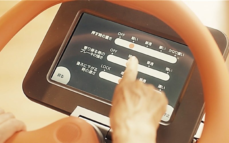タブレット画面上の設定インターフェース。黒い背景に白いスライダーバーと日本語テキスト。指でスライダーを操作する様子。オレンジ色のフレーム付きディスプレイ