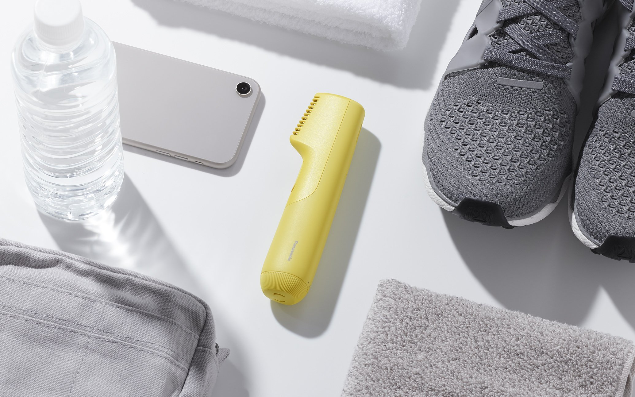 灰色のスマートフォン、黄色のトリマー、透明な水のボトル、白いタオルとバスタオル、グレーのスニーカーが並ぶ白い表面の写真  