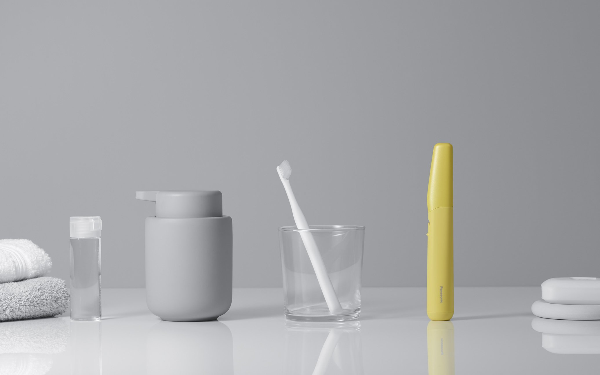 グレーの容器と白の歯ブラシ、黄色の電動歯ブラシ、白いタオルと水のボトルの並ぶ洗面台の写真  