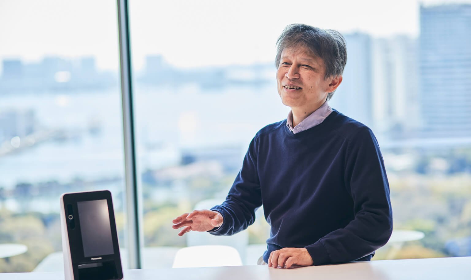 紺色のセーターを着た男性が右手を少し上に上げて話す様子