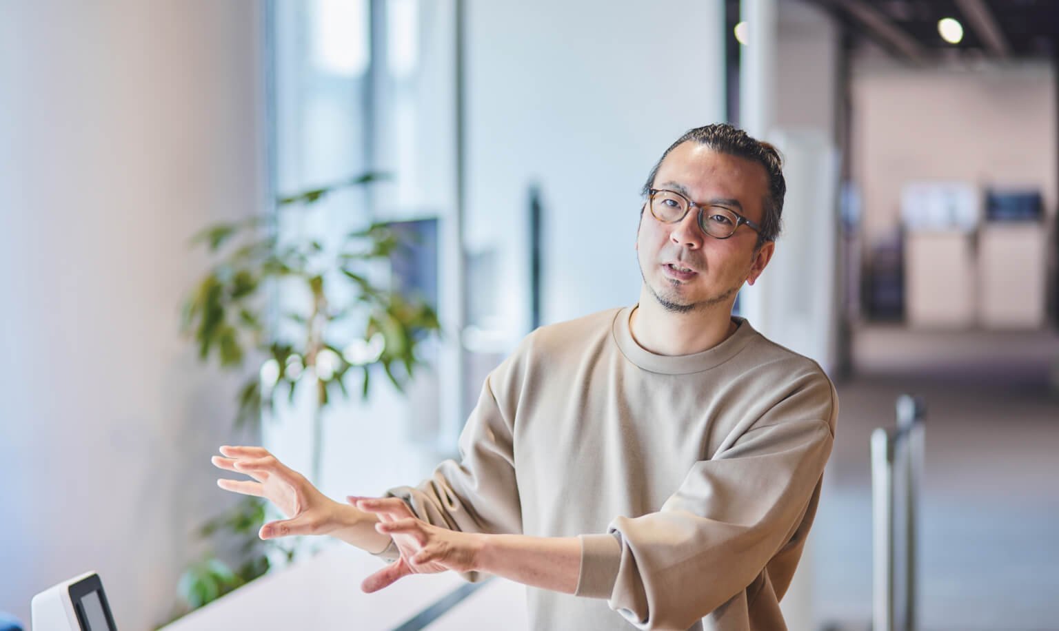 窓辺の室内で両手を前に出しながら話す男性の姿、カジュアルな服装、背景に観葉植物と受付カウンター