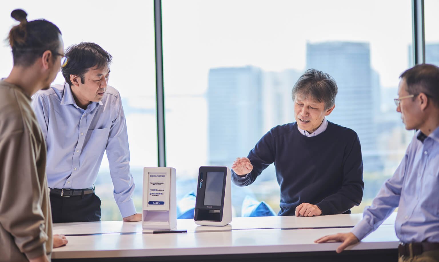 窓際の会議室で四人のビジネスマンが議論、中央に認証付きカードリーダー端末と紙のモックアップモデルが配置