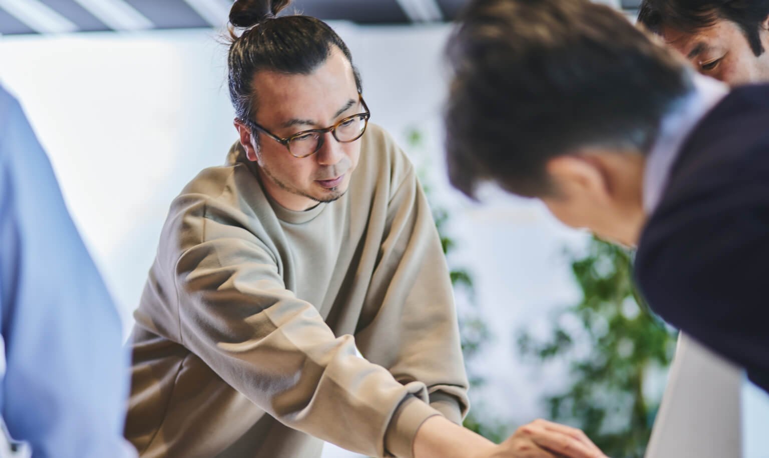 複数人が会議テーブルを囲む様子、中央の男性が手を伸ばして説明している場面