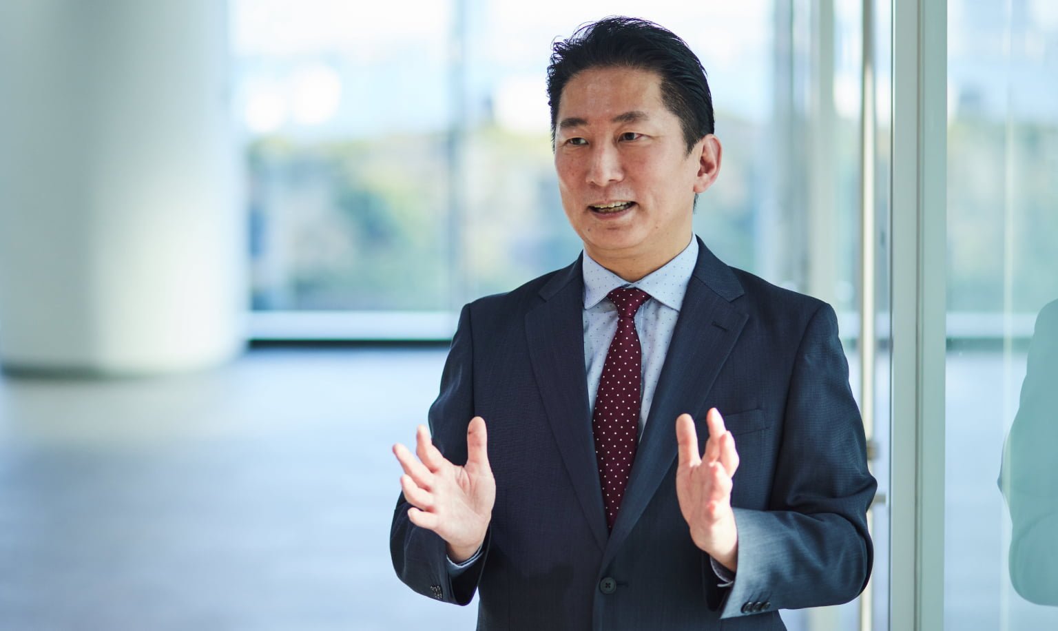 ネイビーのスーツに薄いブルーのシャツ、エンジ色のネクタイをした男性が両手を広げて話している