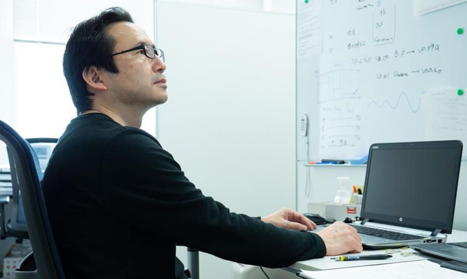 黒い長袖の服を着て眼鏡をかけた男性（森田）が、パソコンを操作しながら正面のホワイトボードを見ている。