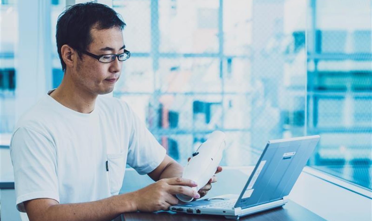 白いTシャツを着た眼鏡をかけた男性（尾形）が、ノート型パソコンの手前で白いミスト発生装置を手にしている