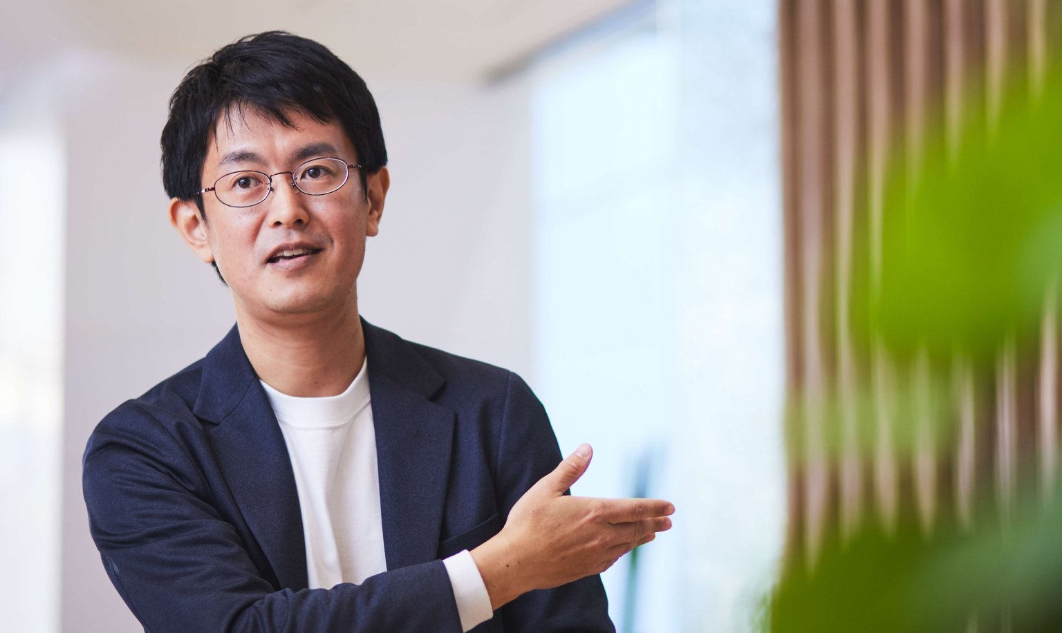 白色シャツに紺色ジャケットを着用しメガネをかけた男性が、右手を前に出して話す姿