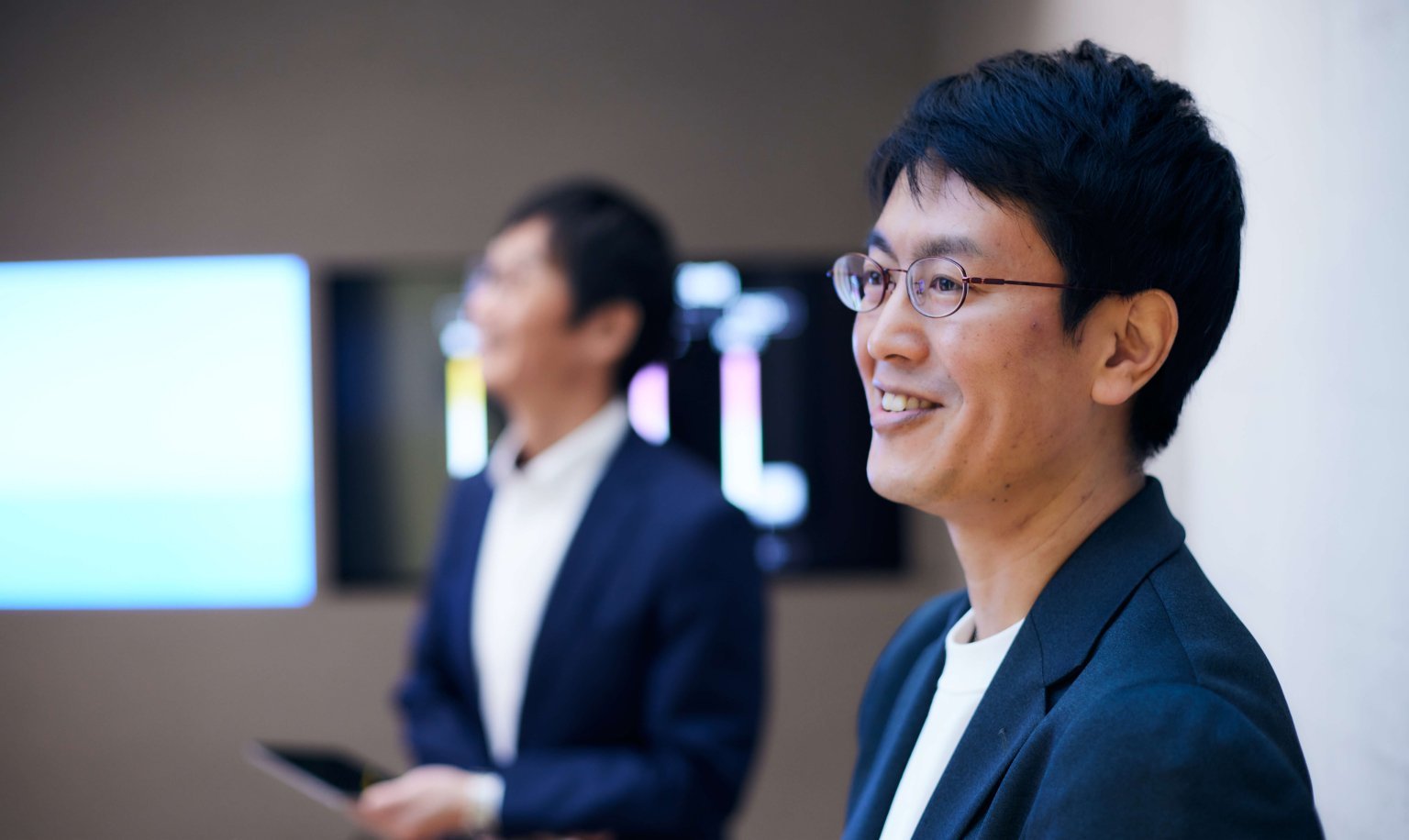 ビジネスマン二人が会議室で座る様子、男性はいずれも白シャツと紺色ジャケット、背景にスクリーンと壁面が見える