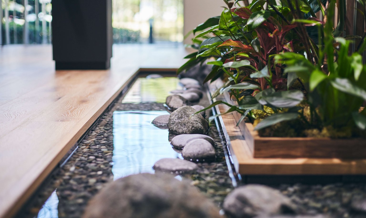 木製の床に沿って配置された石と水が流れる小さな池、池の横に緑色の観葉植物が植えられた木製のプランター