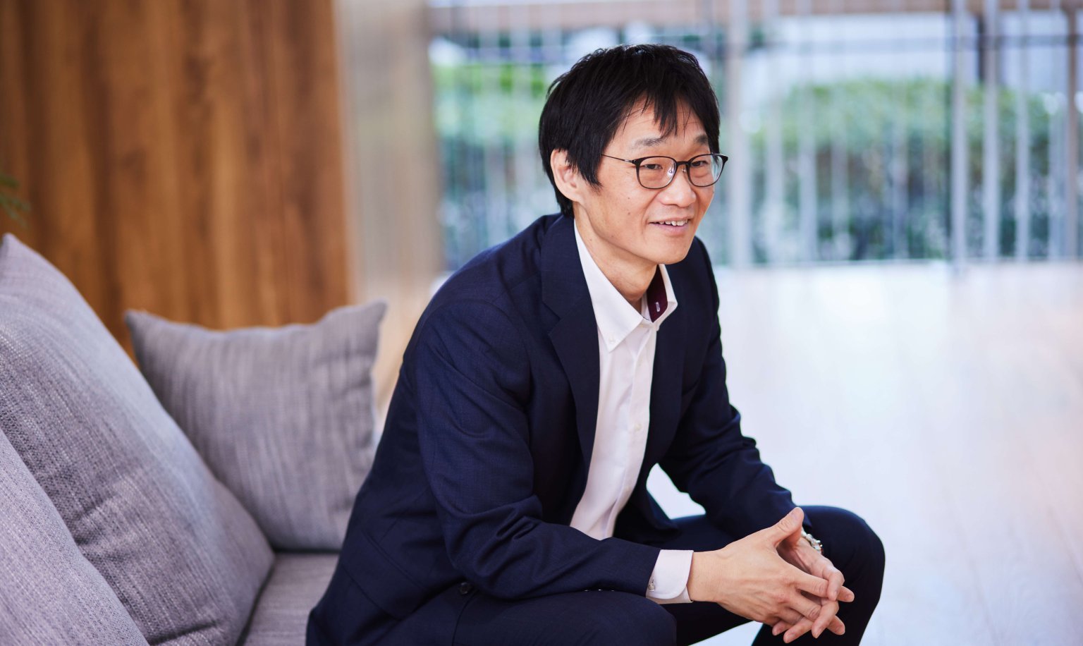 白シャツと紺色ジャケットを着用したメガネの男性が座って手の指を組みソファに座っている姿