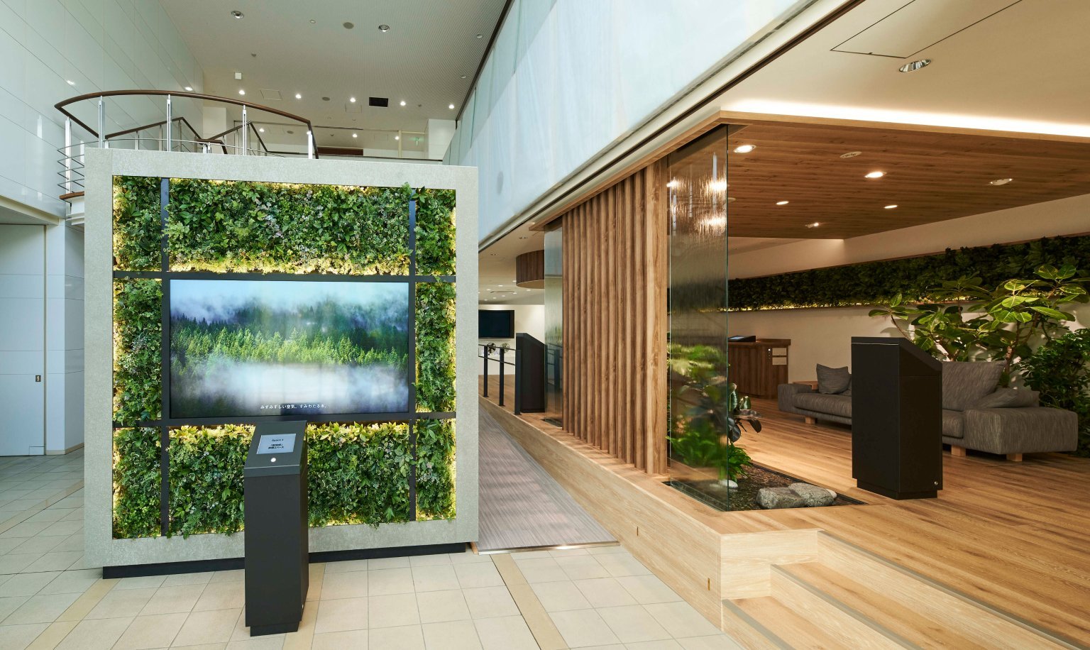 壁面の緑の植物パネルと中央のディスプレイ、木製の仕切りとソファがあるラウンジスペース