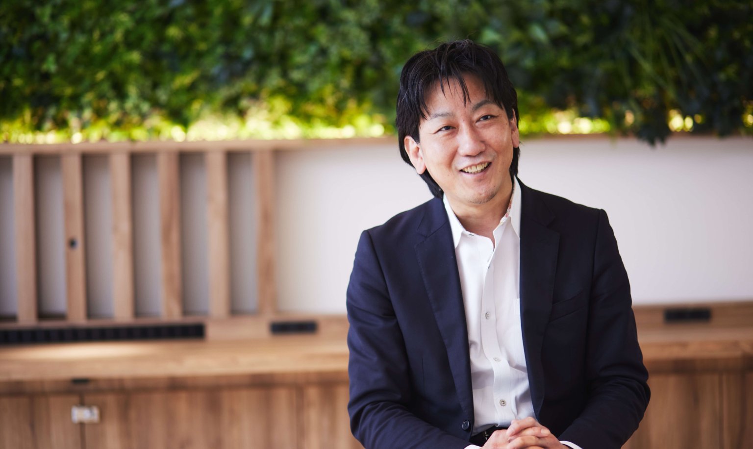 木製のパネルと緑の植栽を背景に、白いシャツに紺色ジャケットを着用した男性が座り、手を組む姿