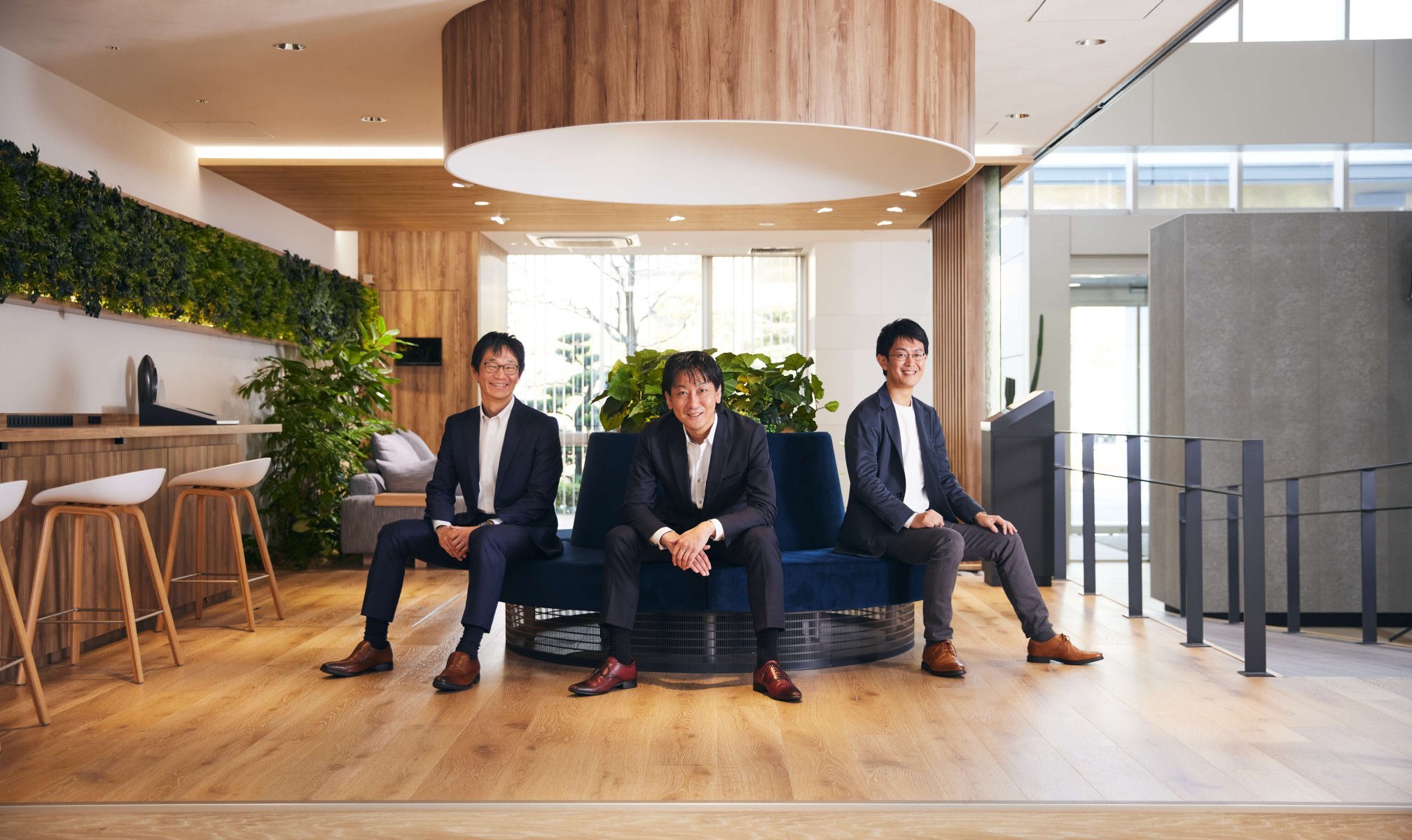 中央に青い円状のソファがあり、その上に大きなフードがある空間に三人のビジネスマンが座る様子、背景に緑の観葉植物と大きな窓、自然光が差し込む