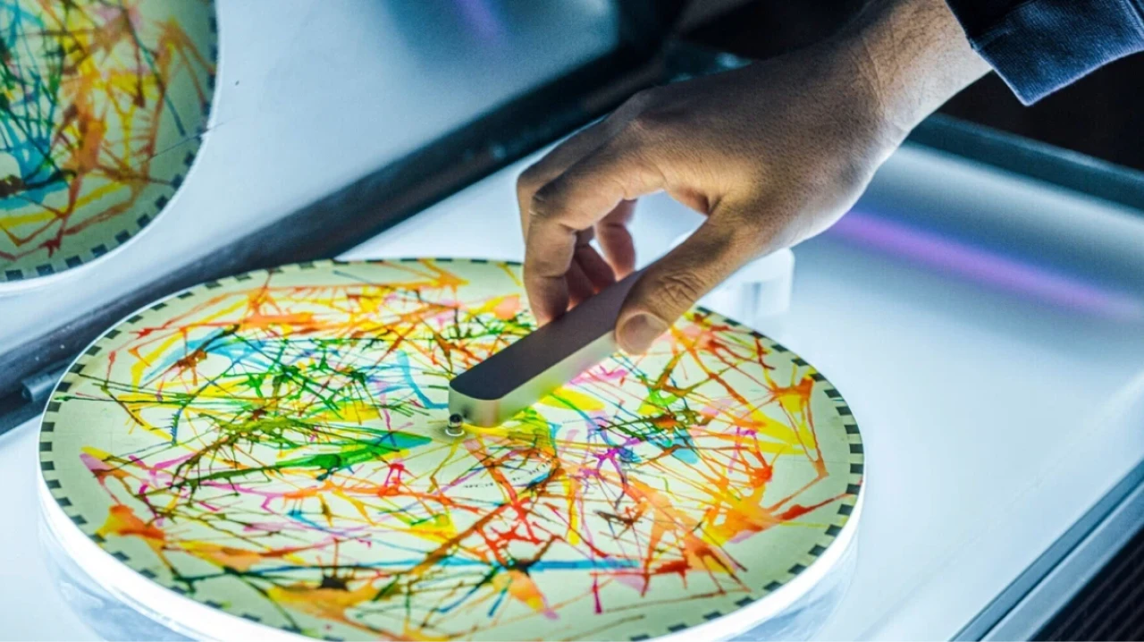 カラフルな線画が描かれた円形のプレートに手を置く様子。手に持たれた金属製の器具がプレートの中心にある。光が当たり、鮮やかな色彩が際立っている。