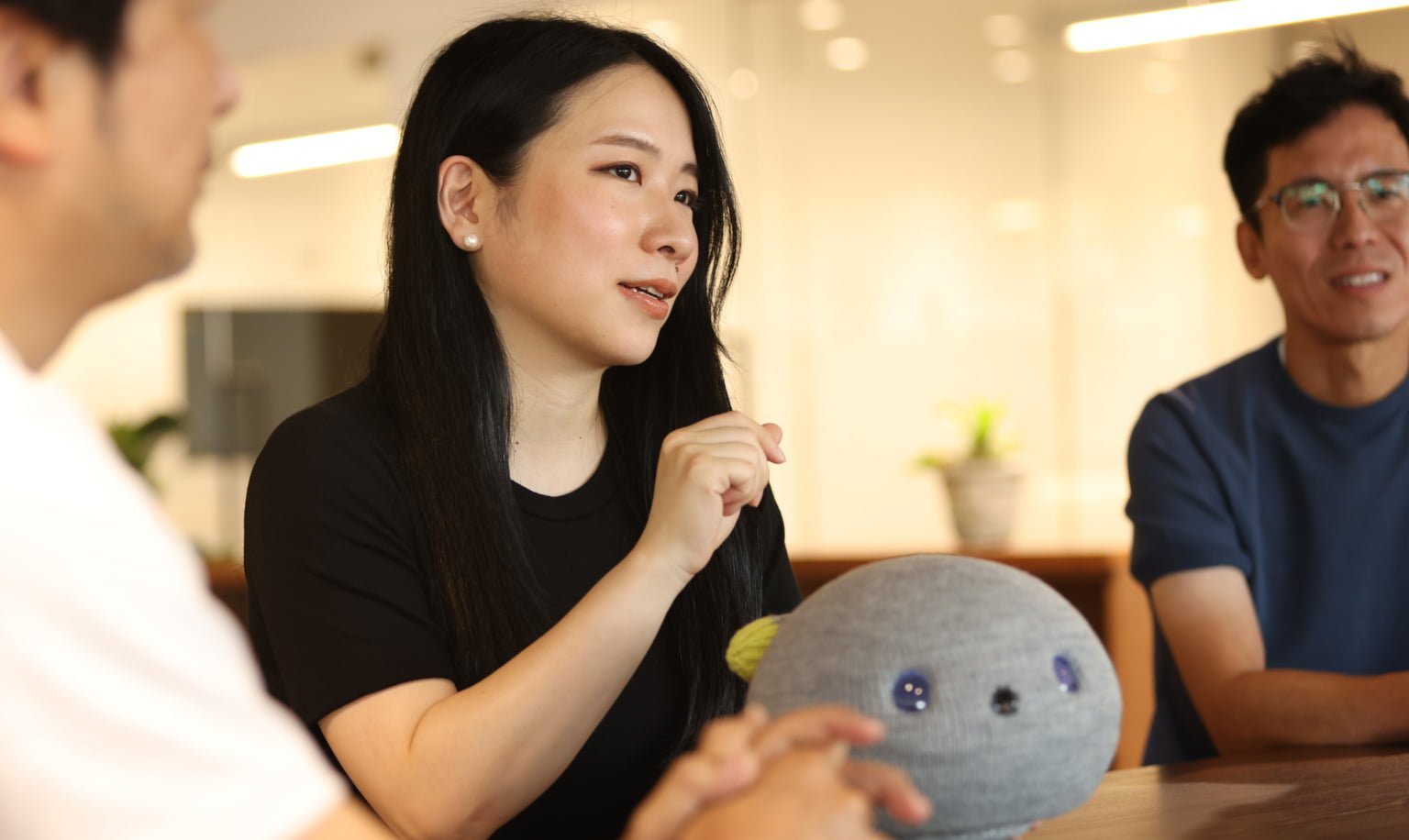 黒い長い髪の女性がテーブルに置かれたニコボを前に置いて話す様子