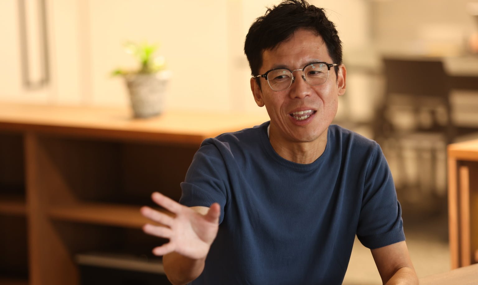 青いシャツを着た男性がテーブルの前で手を上げながら話す様子