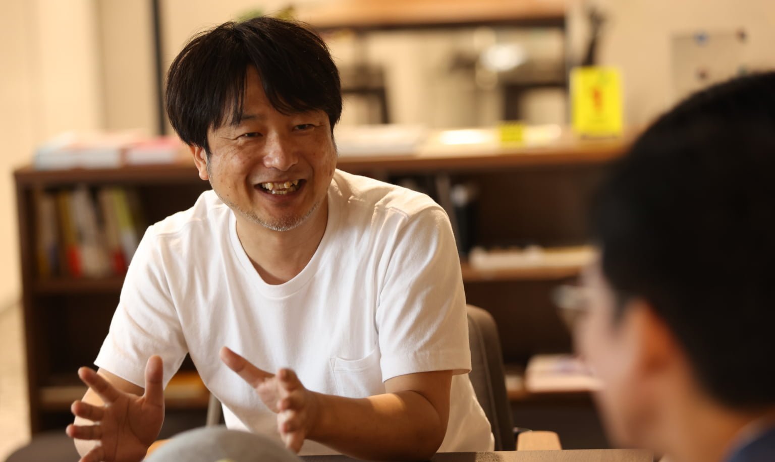 白い Tシャツを着た男性が手を広げながら笑顔で話す様子