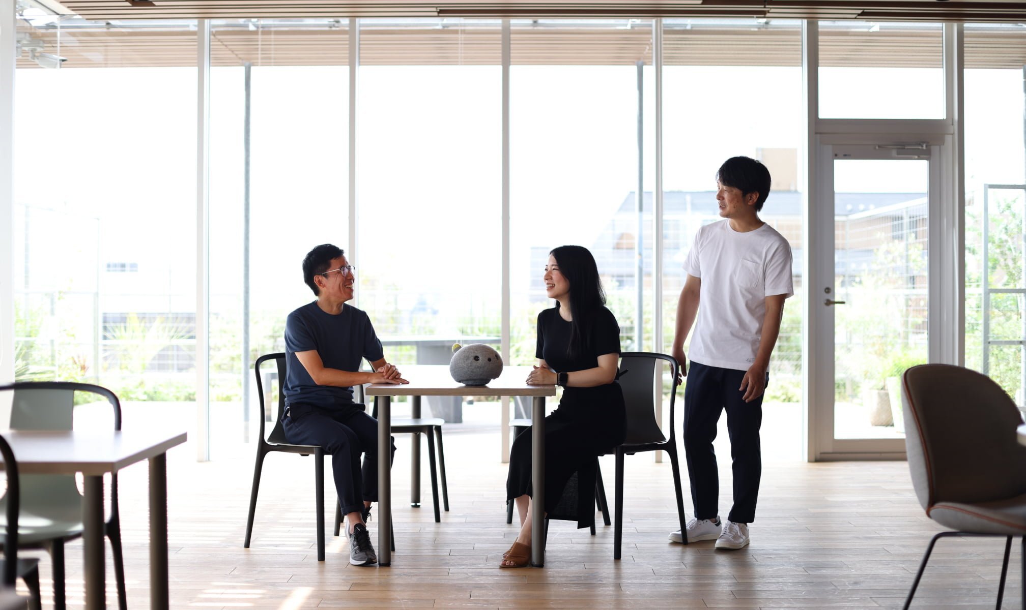 大きな窓のある明るい室内で、三人の若者が会話している様子