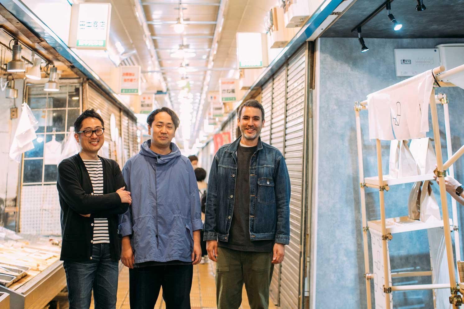 商店街の通路に立ち、横並びでカメラを向く三人の人物