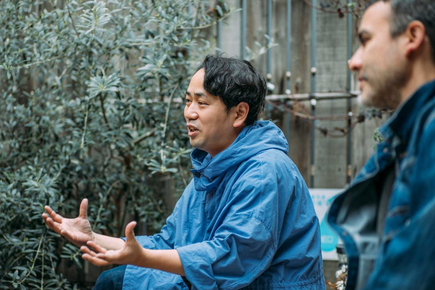 屋外の植栽前で椅子に座り、手振りを交えて話す人物の横顔