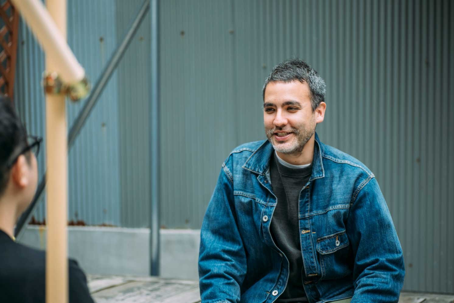 屋外で椅子に座り、正面の人物と話すデニムジャケットの人物