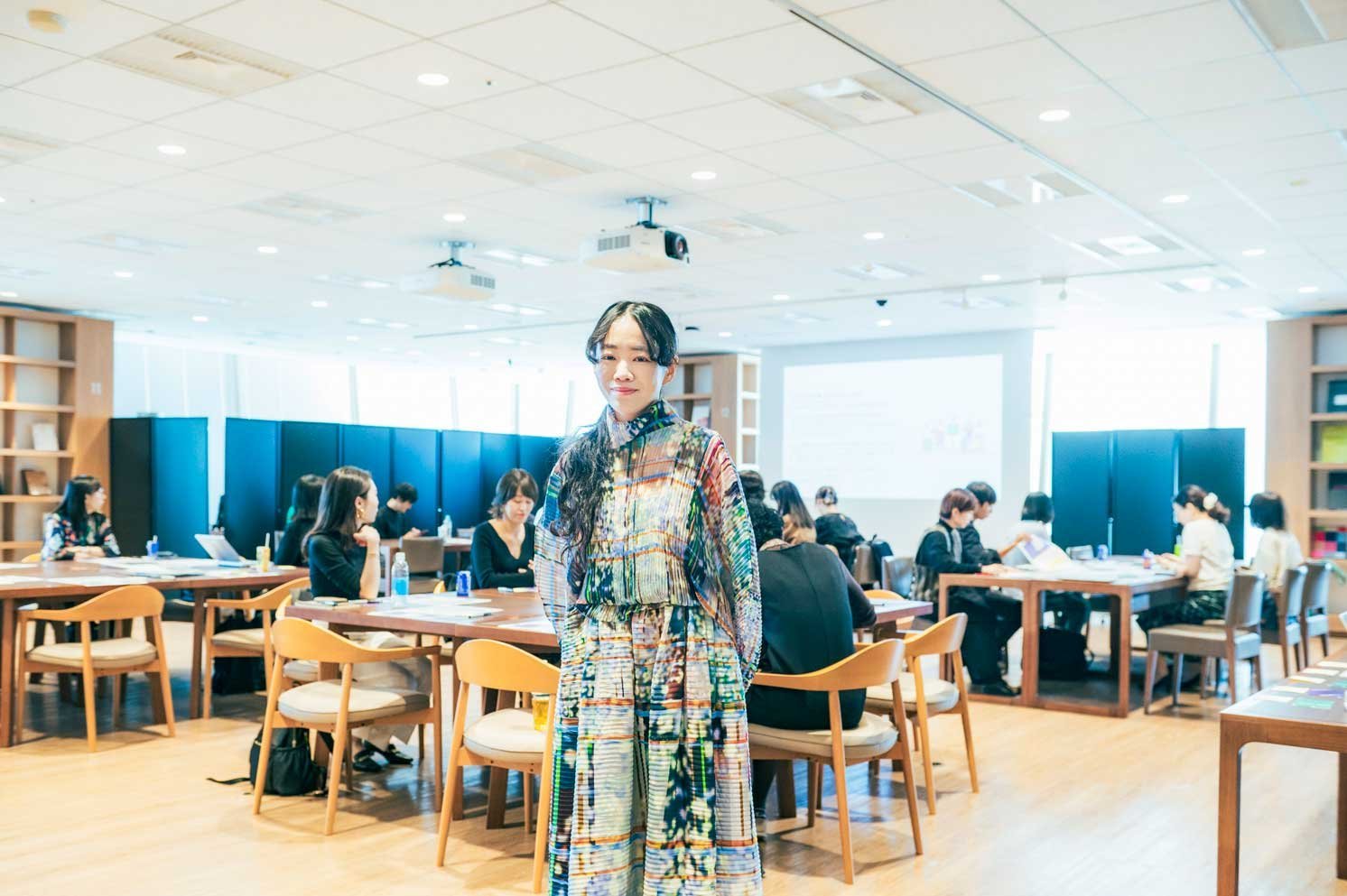 会議室でテーブルを囲み着席する参加者と、中央に立つ人物