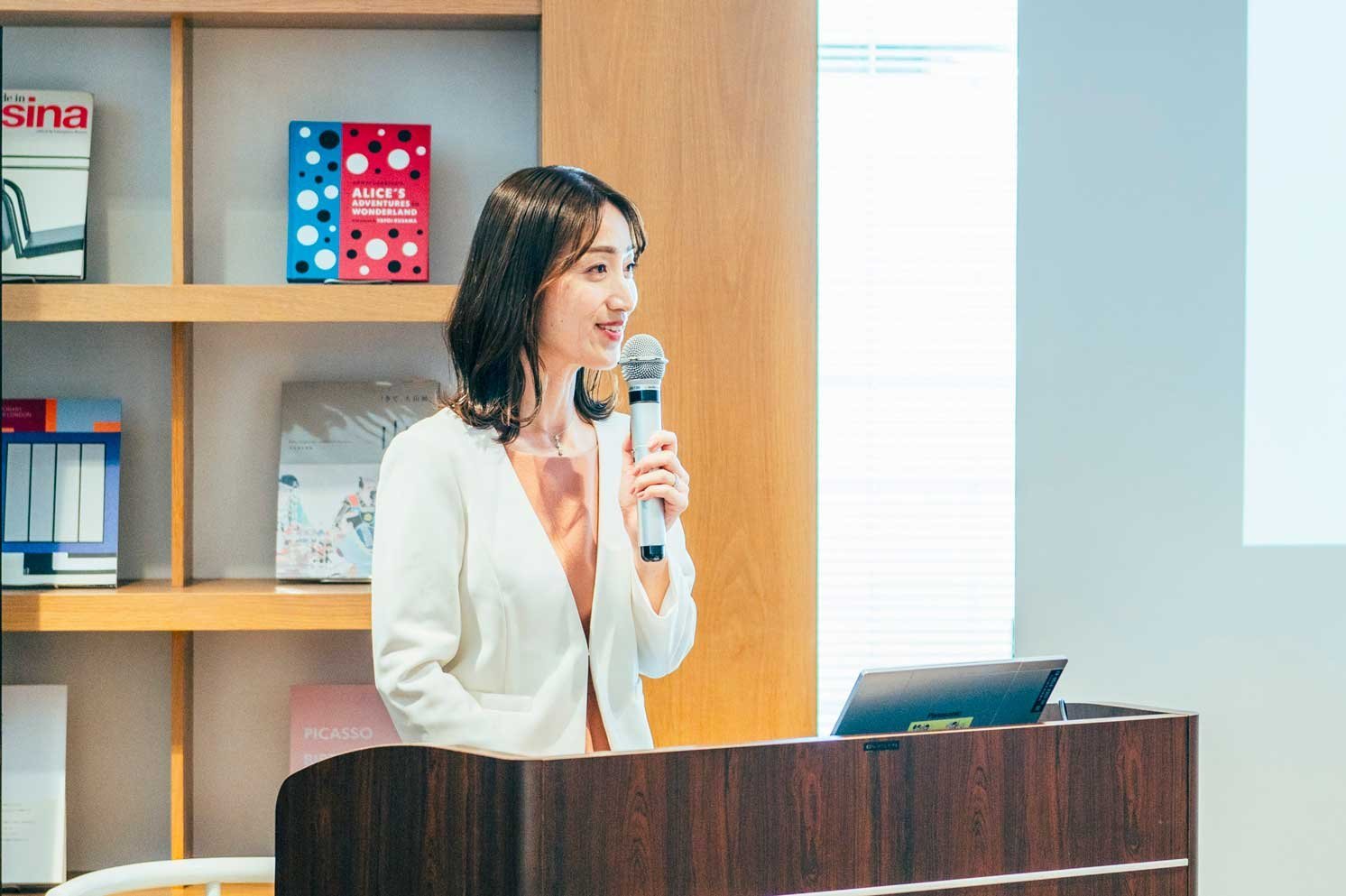演台でマイクを持ち説明する人物と、背後の書棚