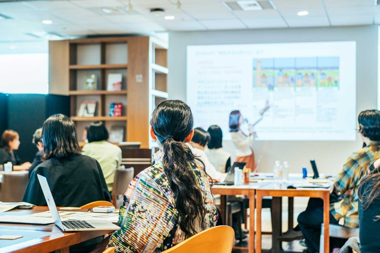 スクリーン前で発表を聞く参加者の後ろ姿と机上の資料