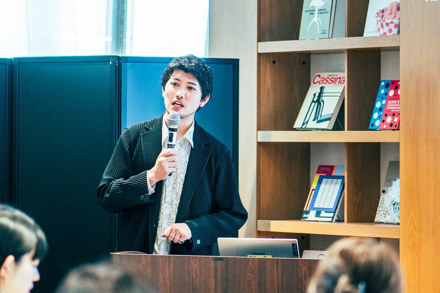 演台でマイクを持ち発表する人物と、書棚に並ぶ書籍