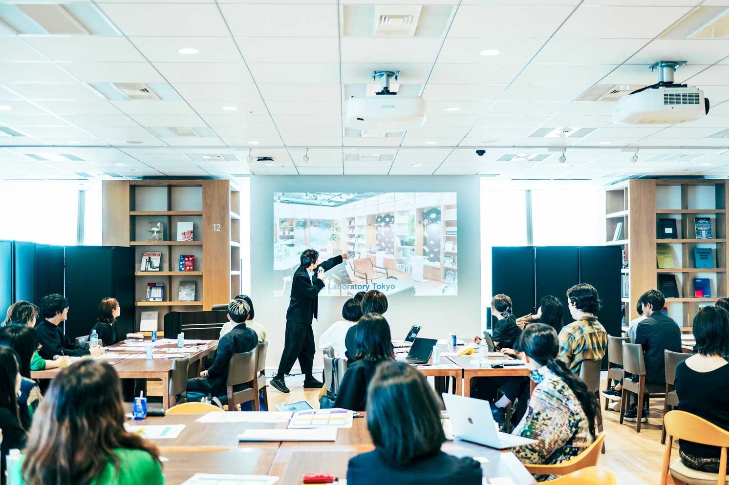 スクリーン前で発表者がスライドを指し示し、着席者が机を囲む会議室