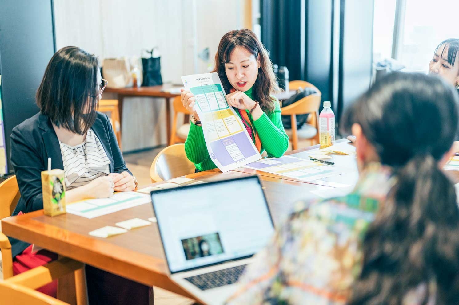 資料を手に説明する人物と、テーブルを囲み着席する複数の人物