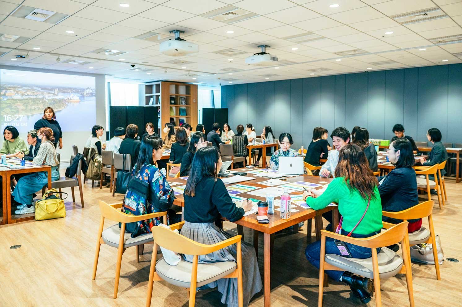 会議室内で複数のテーブルに分かれて着席し、資料を前に話し合う人物