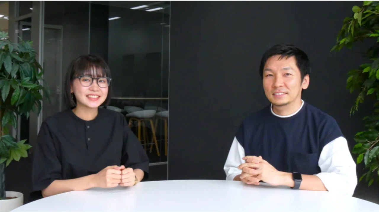 2人の人物が円形のテーブルの前に座っている様子。左側の人物は黒い服を着ており、右側の人物は黒と白の服を着ている。背景には観葉植物とガラスの壁が見える。