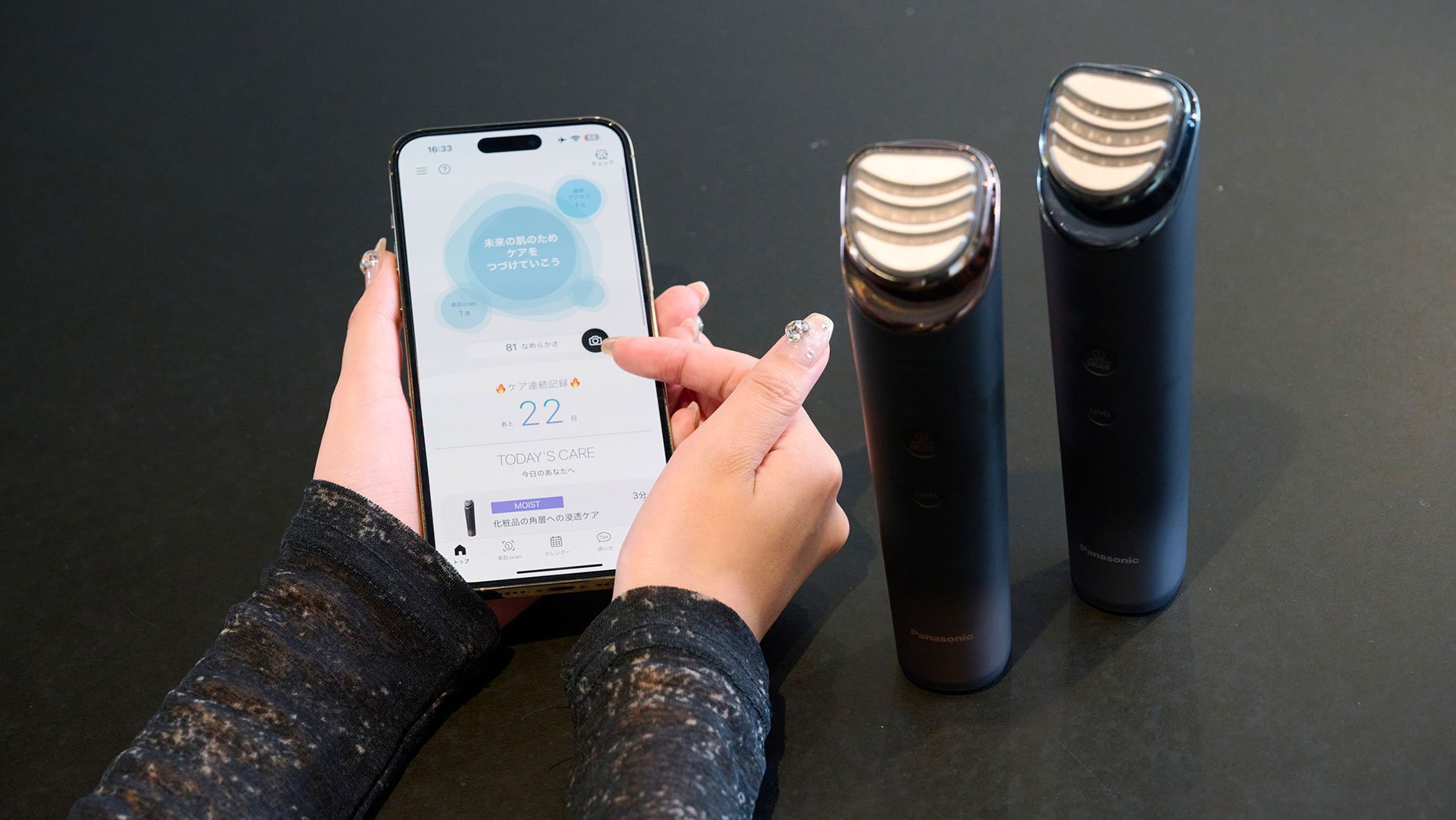 手にスマートフォンを持ち、画面をタッチしている女性の手元が写っている。画面にはスキンケアに関する情報が表示されており、そばにパナソニック製の美容機器が2台置かれている。