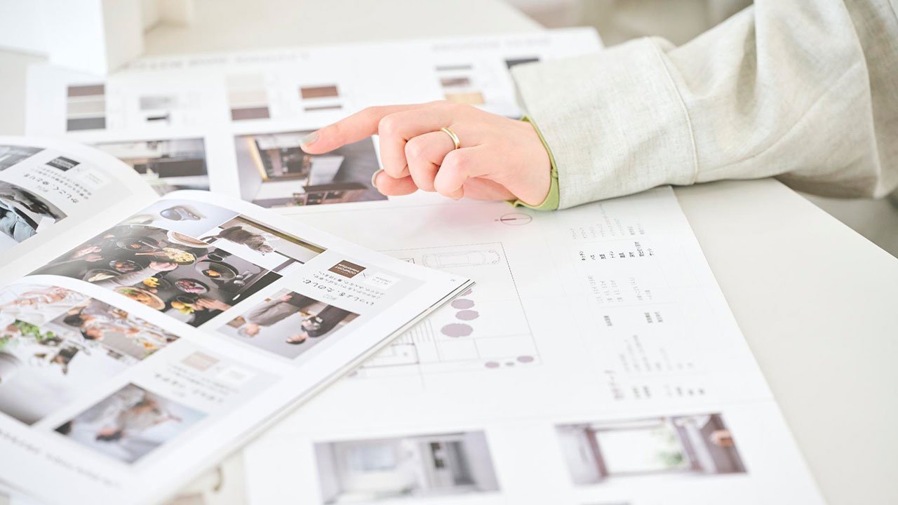 雑誌やカタログを閲覧しながら指差す人の手と腕、 長袖シャツを着用、複数のページと資料が広げられた白いテーブル